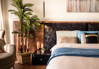 A cozy bedroom featuring a comfortable, well-made bed adorned with multiple pillows in soft and varying hues. A small nightstand with a potted plant sits beside the bed, and an artificial palm plant adds a touch of greenery. Above the bed hangs a painting with abstract tree designs. The warm sunlight streams in through sheer curtains, creating a peaceful and inviting atmosphere.