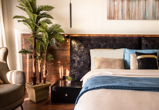 A cozy bedroom featuring a comfortable, well-made bed adorned with multiple pillows in soft and varying hues. A small nightstand with a potted plant sits beside the bed, and an artificial palm plant adds a touch of greenery. Above the bed hangs a painting with abstract tree designs. The warm sunlight streams in through sheer curtains, creating a peaceful and inviting atmosphere.