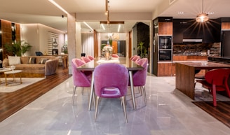 A modern open-plan interior featuring a dining area with a dark wooden table surrounded by pink upholstered chairs, extending to a kitchen and living area. The room is well-lit, with a mix of wooden and marble flooring. The kitchen has dark cabinets accented with a patterned stone backsplash, and there's a stylish light fixture above the table. The living area includes a plush, light-colored sofa, with plants and decorative items enhancing the space.