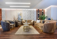 A luxurious living room features plush sofas and armchairs arranged around a central coffee table set with multiple levels. The decor includes elegant throw pillows and a soft area rug on a polished wooden floor. A wall adorned with decorative panels and a large abstract artwork serves as a backdrop. There are plants, a side table with vases, and ambient lighting that enhances the sophisticated ambiance.