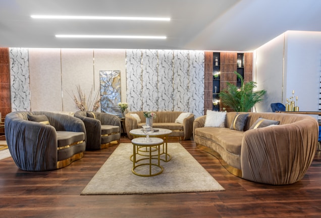 A luxurious living room features plush sofas and armchairs arranged around a central coffee table set with multiple levels. The decor includes elegant throw pillows and a soft area rug on a polished wooden floor. A wall adorned with decorative panels and a large abstract artwork serves as a backdrop. There are plants, a side table with vases, and ambient lighting that enhances the sophisticated ambiance.