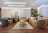 A luxurious living room features plush sofas and armchairs arranged around a central coffee table set with multiple levels. The decor includes elegant throw pillows and a soft area rug on a polished wooden floor. A wall adorned with decorative panels and a large abstract artwork serves as a backdrop. There are plants, a side table with vases, and ambient lighting that enhances the sophisticated ambiance.