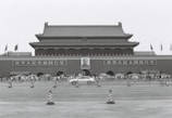A large traditional Chinese gate with a portrait of a leader in the center. Crowds of people stand in front, flanked by signs with Chinese characters. Flags are seen flying above the structure, and a car speeds by in the foreground.