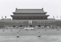 A large traditional Chinese gate with a portrait of a leader in the center. Crowds of people stand in front, flanked by signs with Chinese characters. Flags are seen flying above the structure, and a car speeds by in the foreground.