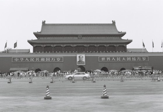 A large traditional Chinese gate with a portrait of a leader in the center. Crowds of people stand in front, flanked by signs with Chinese characters. Flags are seen flying above the structure, and a car speeds by in the foreground.