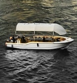 A stylish white boat with a canopy is sailing on textured, dark water. Several people are onboard, one of whom is raising their hand. The boat interior includes tables and chairs, suggesting a relaxed or leisure setting. Ambient lighting highlights the contours of the boat.