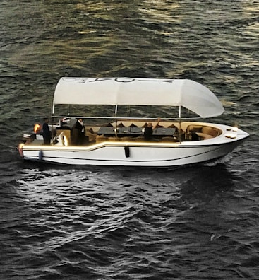 A stylish white boat with a canopy is sailing on textured, dark water. Several people are onboard, one of whom is raising their hand. The boat interior includes tables and chairs, suggesting a relaxed or leisure setting. Ambient lighting highlights the contours of the boat.