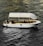 A stylish white boat with a canopy is sailing on textured, dark water. Several people are onboard, one of whom is raising their hand. The boat interior includes tables and chairs, suggesting a relaxed or leisure setting. Ambient lighting highlights the contours of the boat.