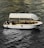 A stylish white boat with a canopy is sailing on textured, dark water. Several people are onboard, one of whom is raising their hand. The boat interior includes tables and chairs, suggesting a relaxed or leisure setting. Ambient lighting highlights the contours of the boat.