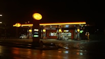A brightly lit gas station at night with a convenience store and fast-food outlet. The forecourt has visible fuel pumps, and there are a couple of cars parked near the fueling area. The ground appears wet, suggesting recent rain, while the yellow-orange lights cast a warm glow on the surroundings.