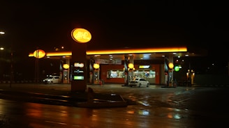 A brightly lit gas station at night with a convenience store and fast-food outlet. The forecourt has visible fuel pumps, and there are a couple of cars parked near the fueling area. The ground appears wet, suggesting recent rain, while the yellow-orange lights cast a warm glow on the surroundings.