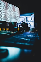 A dimly lit room with a computer setup, featuring a screen displaying abstract line patterns. A mechanical keyboard, headphones, and various computer components can be seen with subdued lighting. Blue and white hues dominate the scene, giving it a futuristic and tech-centric feel.