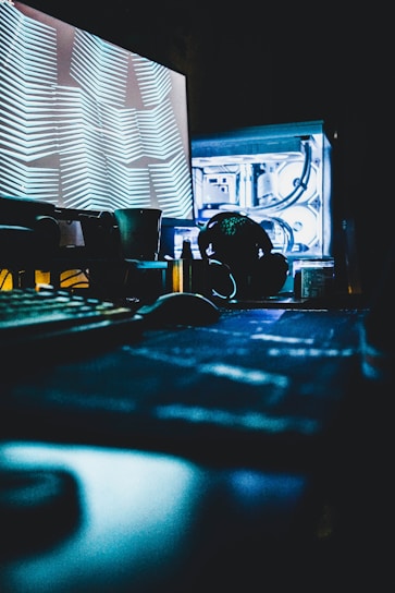 A dimly lit room with a computer setup, featuring a screen displaying abstract line patterns. A mechanical keyboard, headphones, and various computer components can be seen with subdued lighting. Blue and white hues dominate the scene, giving it a futuristic and tech-centric feel.