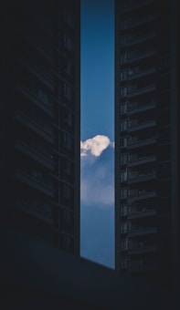 A cloud is visible in the narrow opening between two tall, dark buildings. The sky is clear and blue with the cloud being the focal point against the dark silhouette of the buildings.