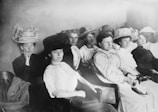 A group of women from the early 20th century seated in a row. They are wearing formal attire typical of the era, including elaborate hats and long dresses. The expressions on their faces are serious and focused, suggesting they might be attending an important event or meeting.