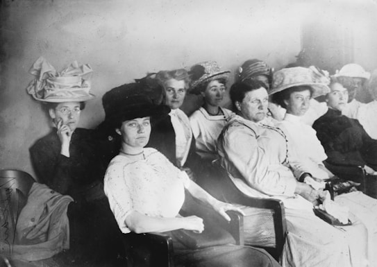 A group of women from the early 20th century seated in a row. They are wearing formal attire typical of the era, including elaborate hats and long dresses. The expressions on their faces are serious and focused, suggesting they might be attending an important event or meeting.