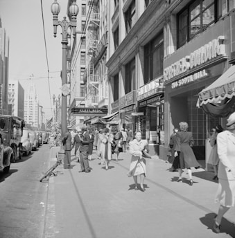 A bustling city street scene from the mid-20th century, showing people in formal attire walking along the sidewalk next to retail stores and buildings. Classic cars are parked along the street, and vintage signage is visible on the storefronts. The architecture is characteristic of early to mid-century urban design, and a few American flags can be seen in the background.