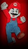 A hand-drawn cartoon character wearing a red hat with an 'M' emblem, red shirt, and blue overalls is shown in a dynamic jumping pose with a raised fist. The background is solid red, emphasizing the character's vibrant colors.