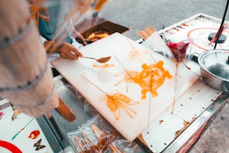 A person is crafting intricate designs using melted sugar on a flat surface, likely at a street food stall. The designs resemble animals and flowers and are being created with a spoon and sticks.