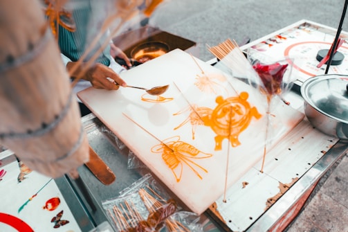 A person is crafting intricate designs using melted sugar on a flat surface, likely at a street food stall. The designs resemble animals and flowers and are being created with a spoon and sticks.