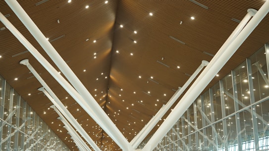 A modern architectural ceiling with wooden paneling and numerous small, bright lights. Large white structural beams converge in a V-shape, and a row of glass windows is visible on the right side.