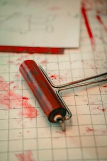 A red ink roller rests on a grid-patterned surface, with splatters of red ink scattered around. Papers with faint markings are visible in the background.