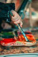 A hand holding a paintbrush dips into a colorful palette filled with bright paints. The surface is splattered with a variety of colors, suggesting an active painting session.