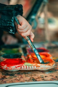 A hand holding a paintbrush dips into a colorful palette filled with bright paints. The surface is splattered with a variety of colors, suggesting an active painting session.