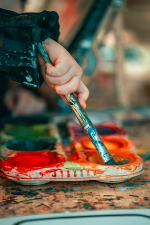 A hand holding a paintbrush dips into a colorful palette filled with bright paints. The surface is splattered with a variety of colors, suggesting an active painting session.