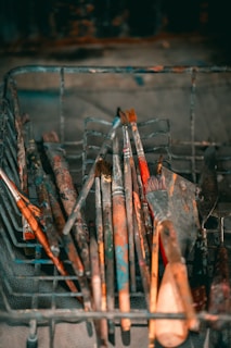 A collection of well-used paintbrushes and putty knives, coated with dried paint, rests in a wire basket. The assortment includes brushes of varying sizes and shapes, displaying hues of red, blue, and brown. The metal basket, set against a blurred background, gives a rustic and artistic feel.