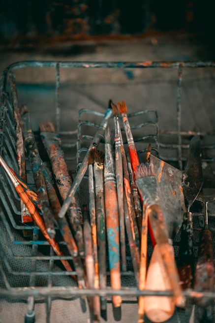 A collection of well-used paintbrushes and putty knives, coated with dried paint, rests in a wire basket. The assortment includes brushes of varying sizes and shapes, displaying hues of red, blue, and brown. The metal basket, set against a blurred background, gives a rustic and artistic feel.