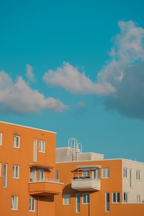 Modern apartment buildings with a bold orange color stand against a clear blue sky. Fluffy white clouds float above, adding a sense of calm and openness. The architecture features clean lines and several balconies, which contribute to a contemporary feel.