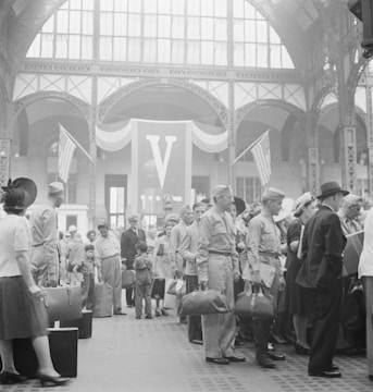 A large group of people gathered inside a grand building with high arched ceilings and large windows. Many are dressed in military uniforms, and some hold luggage. There are American flags and a prominent 'V' banner in the background, suggesting a historical or wartime context. The scene is busy and bustling, with men, women, and children present.