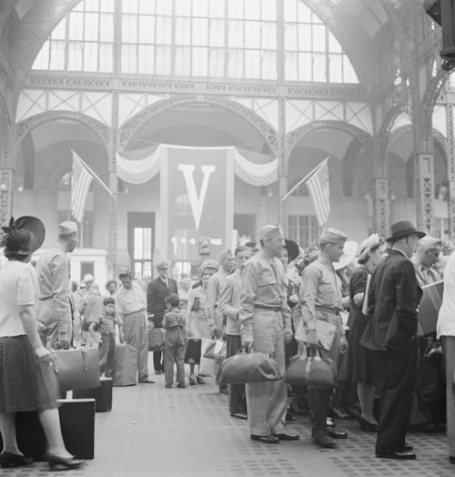 A large group of people gathered inside a grand building with high arched ceilings and large windows. Many are dressed in military uniforms, and some hold luggage. There are American flags and a prominent 'V' banner in the background, suggesting a historical or wartime context. The scene is busy and bustling, with men, women, and children present.