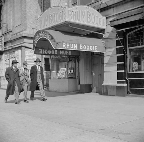 Three men in suits and hats walk past a building with a large marquee sign that reads 'RHUM BOOGIE'. The building has a classic storefront design, and the men appear to be engaged in conversation. In the background, there are posters on the wall, suggesting an urban setting.