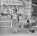 A man stands in front of a storefront filled with signs advertising fresh farm products, including chickens, spare ribs, and lamb. He is dressed in a distinctive outfit adorned with numerous badges and holds a cane and a small bag. To his right, a young boy is sitting on a chair, looking at the camera with a relaxed posture.