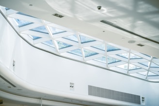 An interior space featuring a modern architectural ceiling design with a large skylight composed of triangular glass panels. The ceiling is primarily white, with a clean and minimalist appearance. Sunlight filters through the glass, casting geometric shadows on the surfaces below.