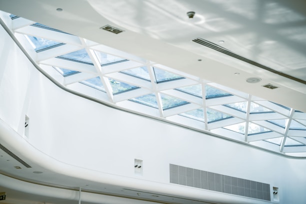 An interior space featuring a modern architectural ceiling design with a large skylight composed of triangular glass panels. The ceiling is primarily white, with a clean and minimalist appearance. Sunlight filters through the glass, casting geometric shadows on the surfaces below.