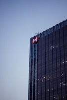 A tall, modern glass skyscraper with a geometric red and white logo on its corner, set against a clear evening sky. The building's reflective surface captures subtle ambient light.
