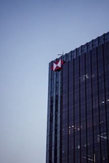 A tall, modern glass skyscraper with a geometric red and white logo on its corner, set against a clear evening sky. The building's reflective surface captures subtle ambient light.