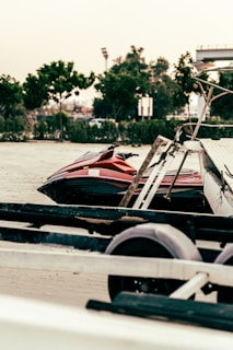 A red jet ski is parked on a sandy area next to some trailers. In the background, there are trees and a partially visible structure, possibly a bridge or building.