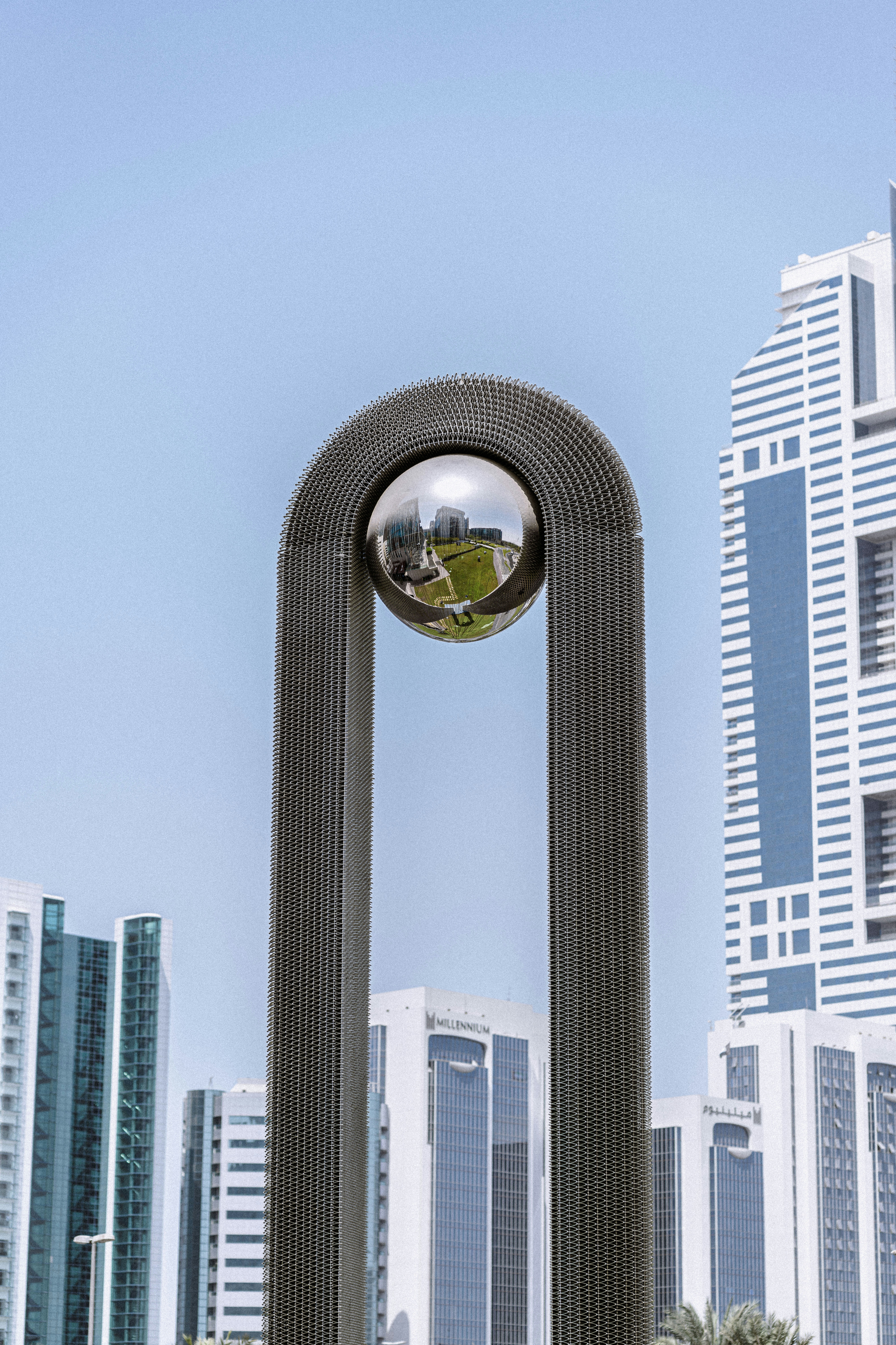 Sculptural installation featuring a reflective orb surrounded by a curved metallic frame, with modern skyscrapers in the background, symbolizing technological advancement in urban settings.