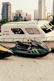 A jet ski with visible branding and green stripes is parked on a sandy area beside a large, worn-out boat. Skyscrapers and city buildings are present in the distant background, providing an urban setting.