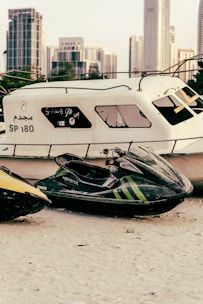 A jet ski with visible branding and green stripes is parked on a sandy area beside a large, worn-out boat. Skyscrapers and city buildings are present in the distant background, providing an urban setting.