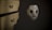 A white mask adorned with black crosses and stitches covers a figure in a dark, hooded garment. It sits next to a wooden dresser with ornate, vintage-style drawer handles. The lighting is dim, creating a mysterious and eerie atmosphere.