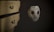 A white mask adorned with black crosses and stitches covers a figure in a dark, hooded garment. It sits next to a wooden dresser with ornate, vintage-style drawer handles. The lighting is dim, creating a mysterious and eerie atmosphere.