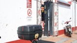 A small motorbike is parked against a red and white wall, next to a gate with decorative metalwork. A 'No Smoking' sign in multiple languages is visible. There is a large black box attached to the back of the motorbike.