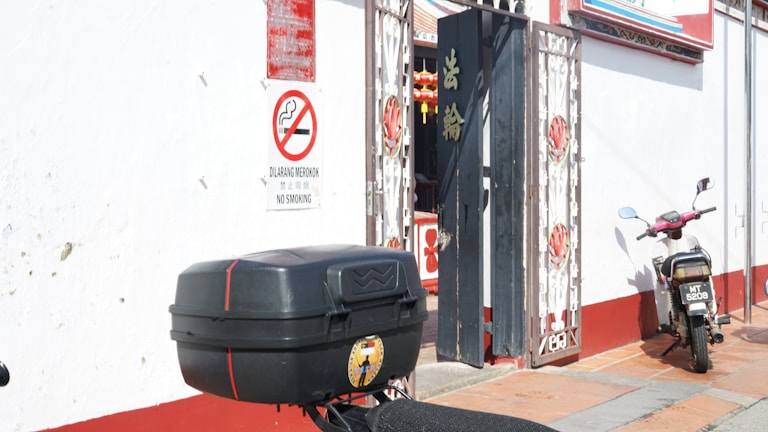 A small motorbike is parked against a red and white wall, next to a gate with decorative metalwork. A 'No Smoking' sign in multiple languages is visible. There is a large black box attached to the back of the motorbike.