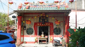 A colorful, traditional Chinese temple adorned with red lanterns, ornate decorations, and intricate architectural details, including a decorative roof with dragon sculptures. In front of the temple are a blue car and a motorcycle, alongside some potted plants. The temple is set against an urban backdrop with nearby buildings visible.