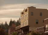 A row of rustic buildings with a prominent sign for a coffee shop offering breakfast and lunch. The architecture features brick and stucco facades with a backdrop of tall pine trees under a partly cloudy sky.